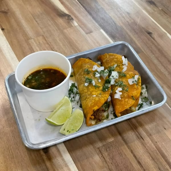 Two birria tacos with cilantro, onions, lime wedges, and a cup of consommé on a metal tray.